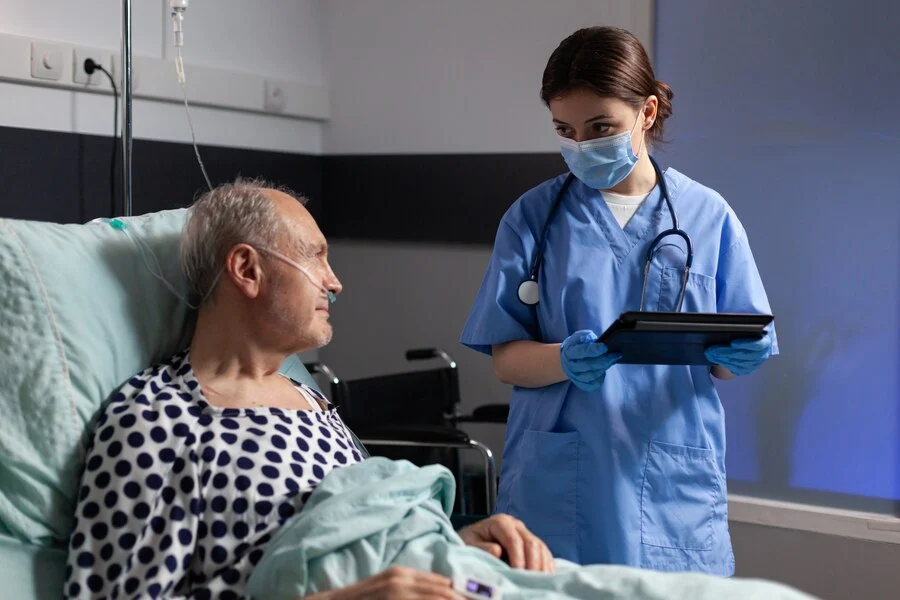 A nurse holding a tablet beside a man in a hospital bed give the urgent care services.
